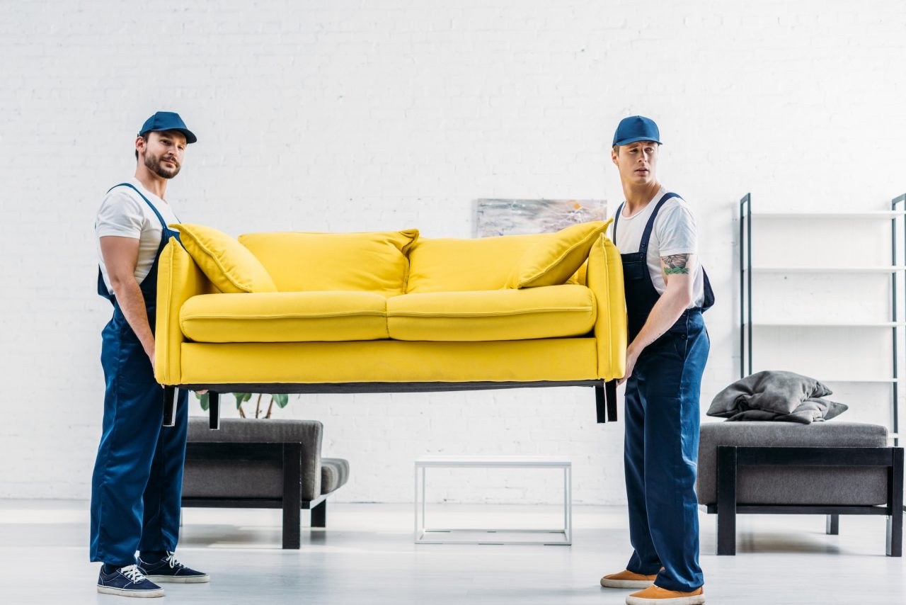 Expert Removals - Two men expertly carrying a yellow couch into a room.
