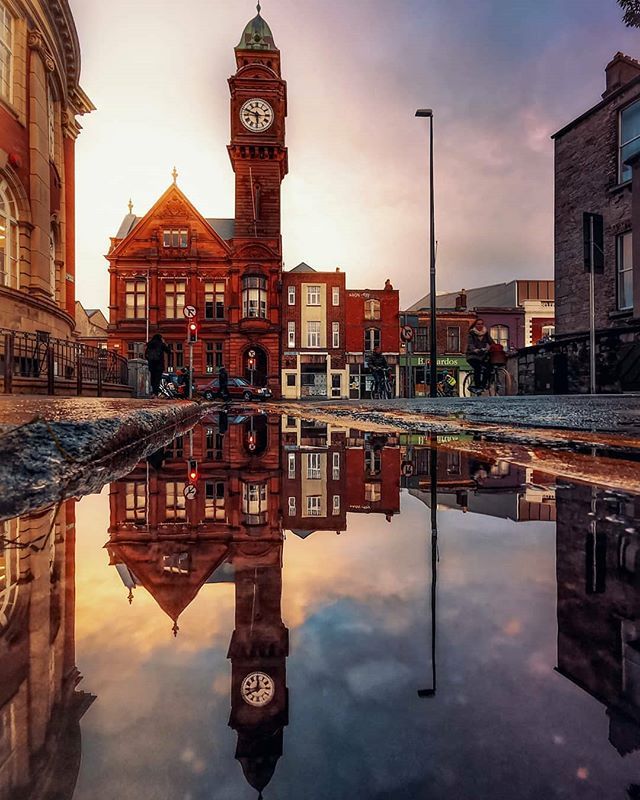 An Expert Removals truck passes by, causing ripples in a puddle that beautifully reflects a nearby clock tower.